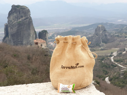 A TerraNut burlap drawstring gift sack and a raw nut snack bar resting on a stone wall, with a scenic mountain landscape and historic rock formations in the background.
