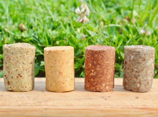 Four different flavors of TerraNut raw, cold-pressed snack bars standing upright in a row on a wooden board with green grass in the background.