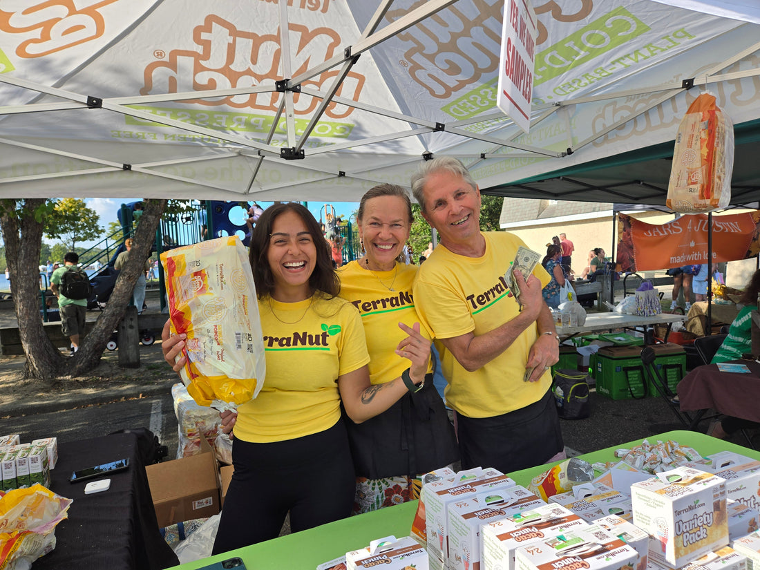 Ula, Ed, and the TerraNut team sampling Nut Punch bars and POP popcorn at a farmers market tent in a park, with a playground in the background