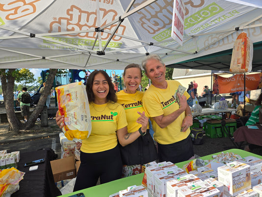 Ula, Ed, and the TerraNut team sampling Nut Punch bars and POP popcorn at a farmers market tent in a park, with a playground in the background