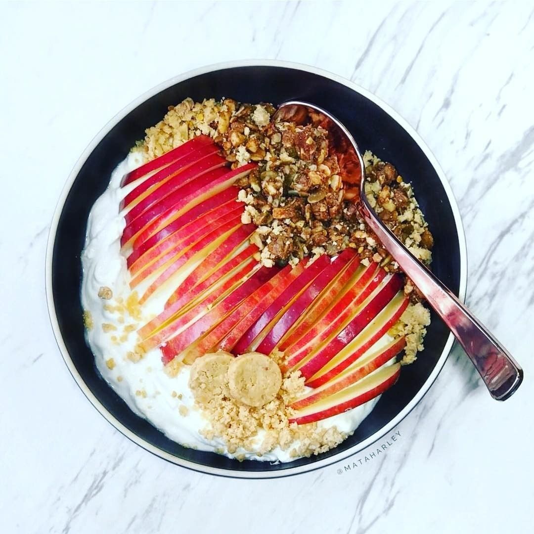 A beautifully arranged breakfast bowl on a marble surface featuring thin apple slices, creamy yogurt, and crumbled TerraNut Pnut Punch with two whole cold-pressed nut cylinders.