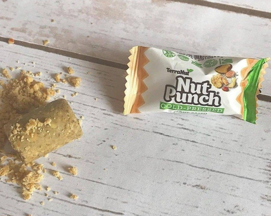 A wrapped TerraNut Nut Punch bar alongside a bite-sized cold-pressed cylinder and crumbs on a white wooden surface, showcasing the authentic texture.