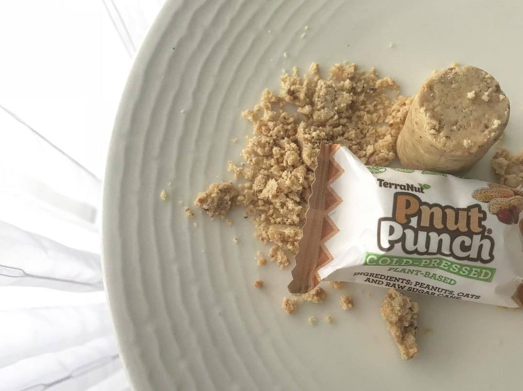 Wrapped TerraNut Pnut Punch cold-pressed snack bar with a cut snack bite and crumbs on a white plate.